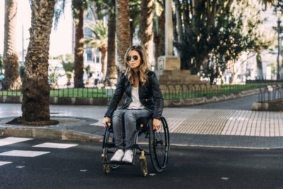 Küschall Ultra-Light - ung kvindelig kørestolsbruger kryfser vejen. Palmer og sol